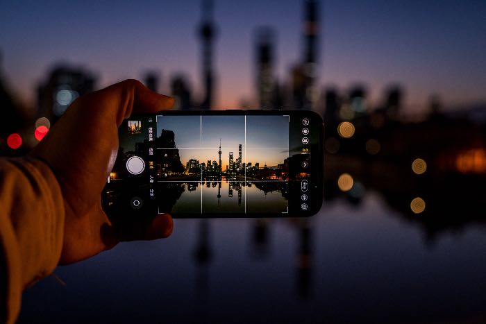 "John's not so sure about Jesus (Advent 3A)" - a photo of a person taking a picture of a cityscape with his phone, with the city image from the phone in focus.