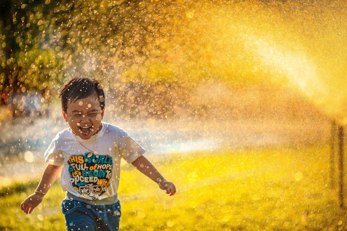 "Baptism Redux (Epiphany 2A)" - a photo of a boy running through a sprinkler, smiling big