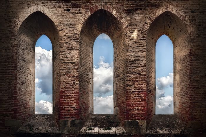 a photo of three tall windows, clouds are visible through them. It appears to be from a very old church, and the clouds make it appear to be around us.