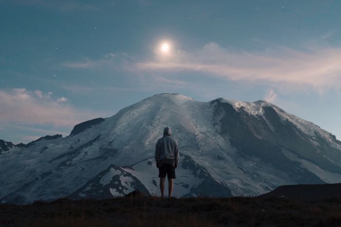 a photo of a person standing in front of a mountain at dusk, the moon, glowing straight above.