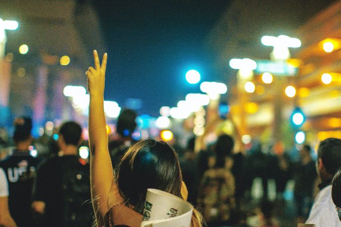 a photo of people marching in the street; a person in the foreground has their hand up in a peace sign