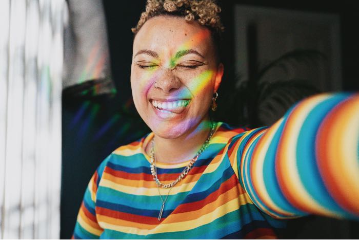 a selfie photo of a person in a rainbow shirt, eyes-closed, big smile, tongue out a little, to the side, with light creating a rainbow on their face.