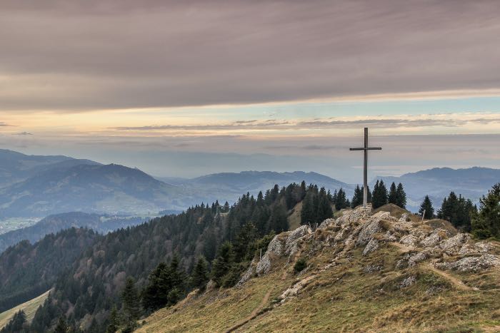 a photo of a cross on a mountaintop, with evergreen trees below