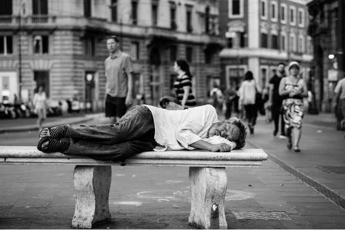 a photo in black and white of a man lying on a bench, sleeping, in the midst of a city with people walking all around