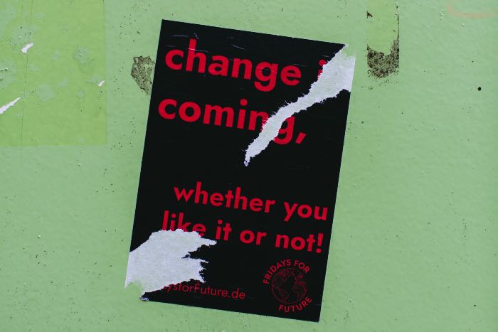 a photo of a black poster on a green wall, torn, as if someone tried to remove it from two different corners. It reads: "change is coming, whether you like it or not!"