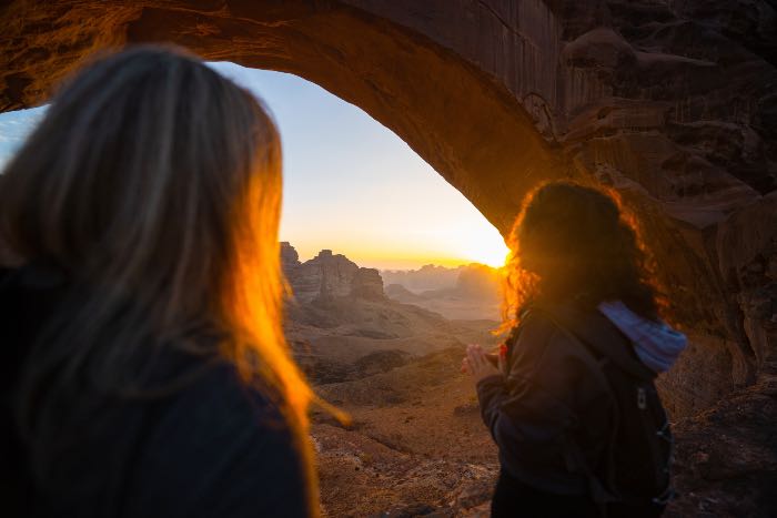 a photo of two women standing near each other, under an arch, seeing the sun setting, peaking out of the corner of the archway.