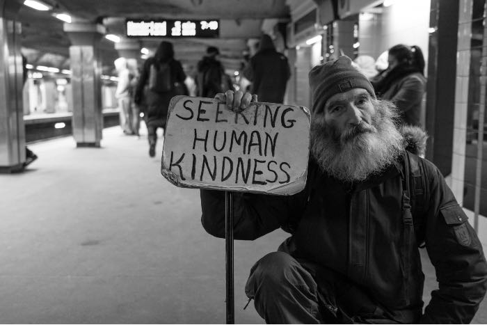 a photo of a man with a beard, kneeling on a subway platform, holding a cardboard sign which reads: "seeking human kindness"