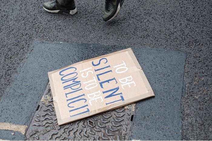 a photo of a city street, a cardboard sign on the sidewalk reads "to be silent is to be complicit".