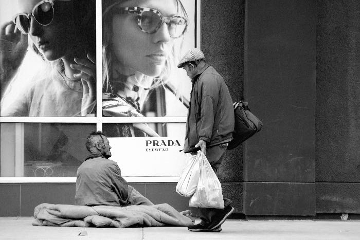 a photo of a person talking to a person sitting on the pavement in front of a Prada billboard.