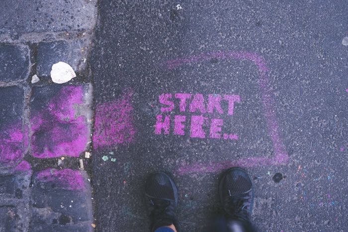 a photo of a street, feet standing before the words painted in purple: "START HERE"