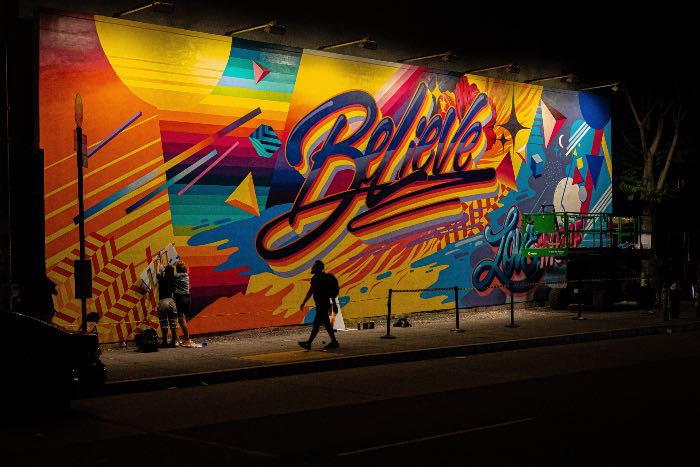a photo of a person walking in front of a colorful mural that has a word in bold cursive: Believe
