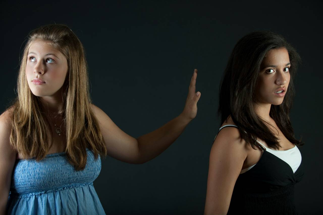 a photo of two young women, one with her back to the other, and one with her hand up and looking away