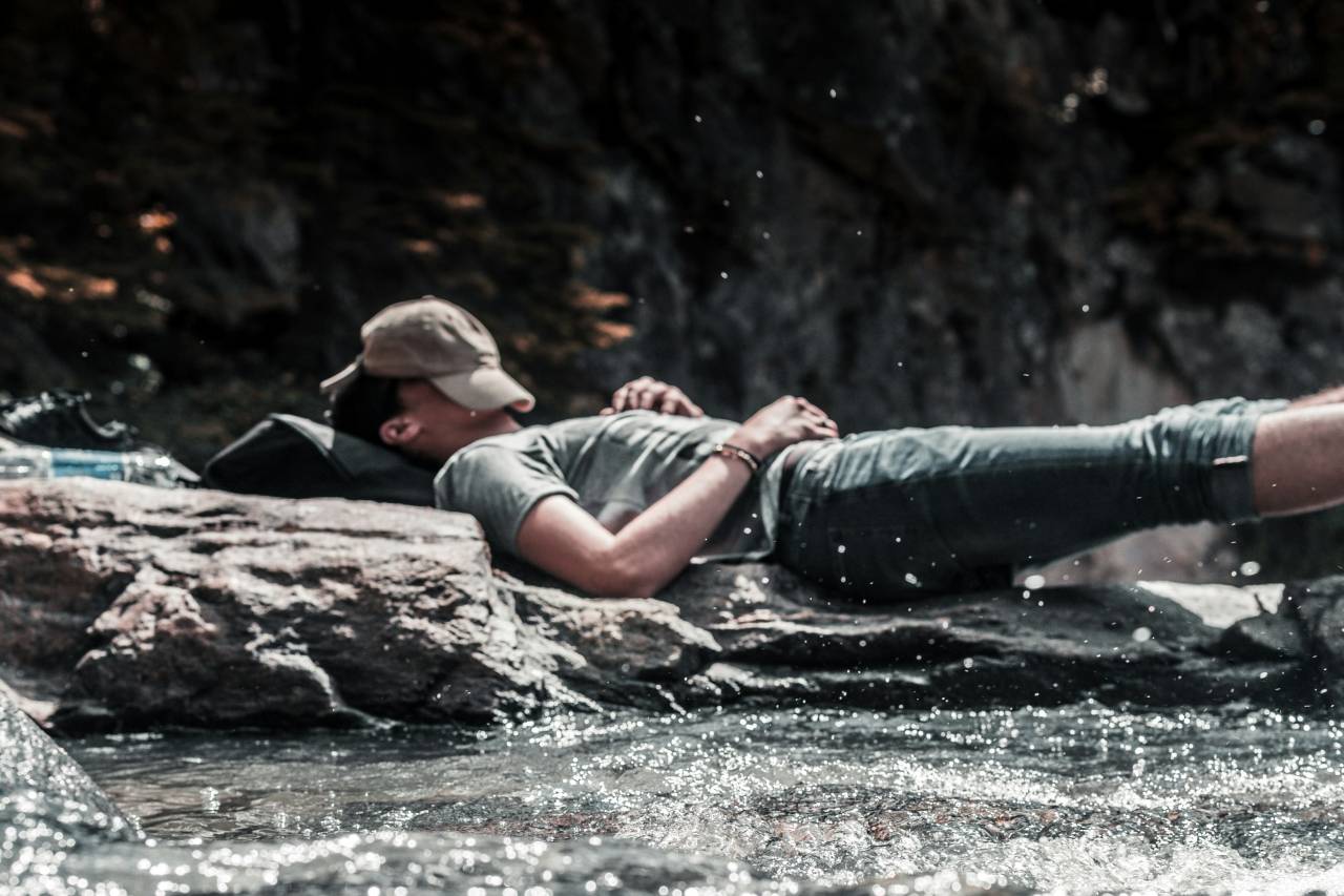 a photo of a young man sleeping by a river bed, with his hat over his face