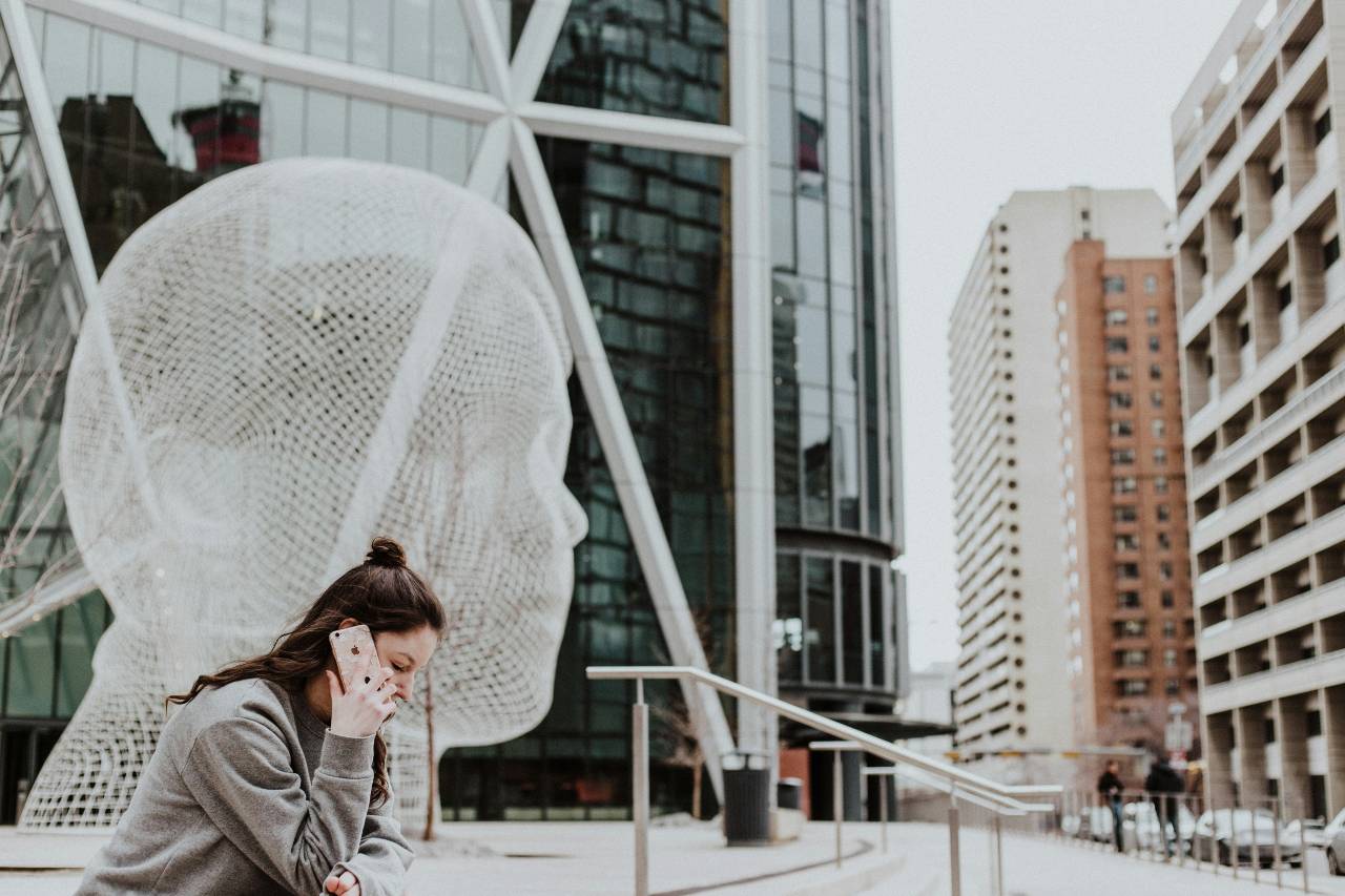 A person on the phone, sitting on the steps of a big urban building, a sculpture of a giant translucent head behind them.