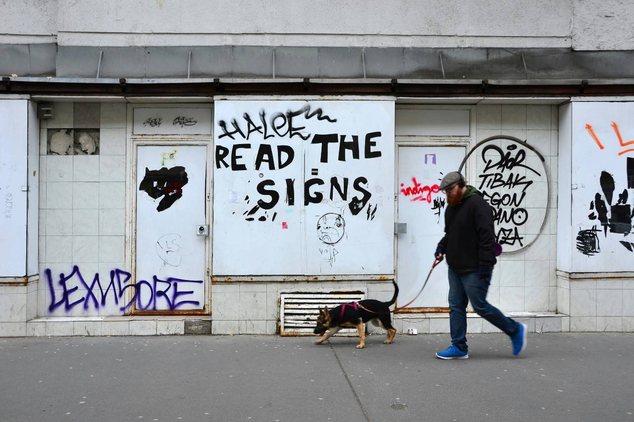 a photo of a person walking his dog, graffiti behind him says "read the signs"