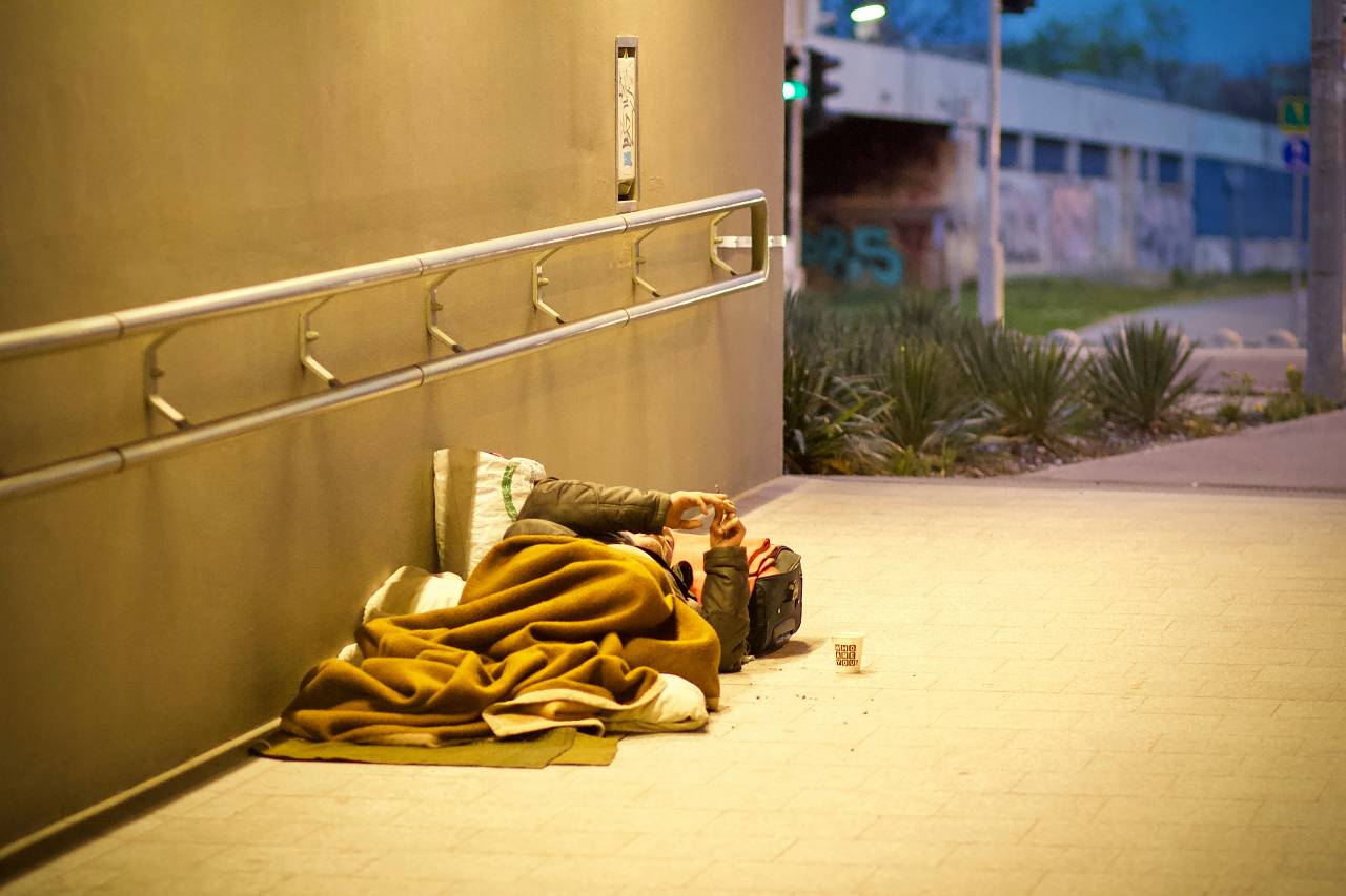 a photo of a person laying against a wall in a public walkway, wrapped in a blanket